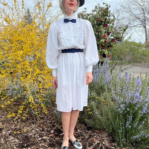 K&ouml;nnte beinhalten: Eine Frau in einem wei&szlig;en Kleid mit einem schwarzen G&uuml;rtel und einem schwarzen Hut mit einer blauen Schleife. Sie steht vor einem Busch mit gelben Bl&uuml;ten.