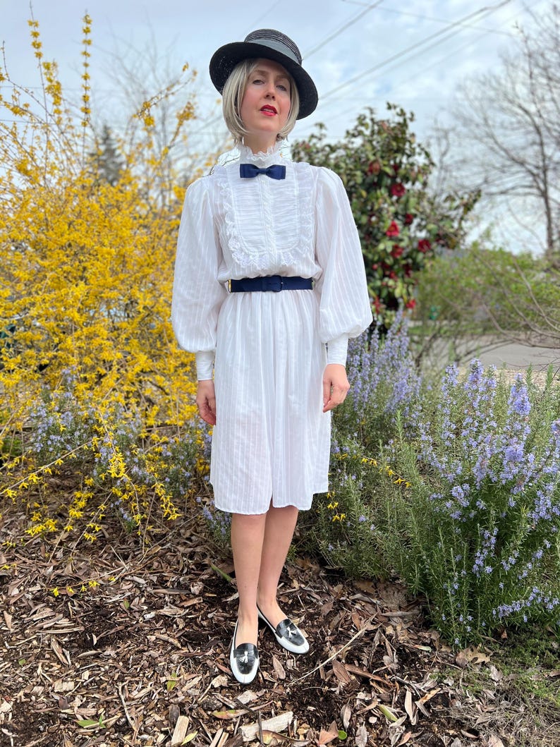 K&ouml;nnte beinhalten: Eine Frau in einem wei&szlig;en Kleid mit einem schwarzen G&uuml;rtel und einem schwarzen Hut mit einer blauen Schleife. Sie steht vor einem Busch mit gelben Bl&uuml;ten.