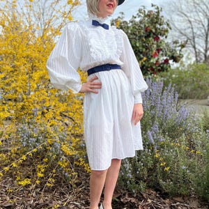 K&ouml;nnte beinhalten: Ein wei&szlig;es Kleid mit einem R&uuml;schenkragen und einer blauen Schleife. Das Kleid hat ein tailliertes Oberteil und einen vollen Rock. Das Model tr&auml;gt einen schwarzen Hut und schwarz-wei&szlig;e Schuhe.