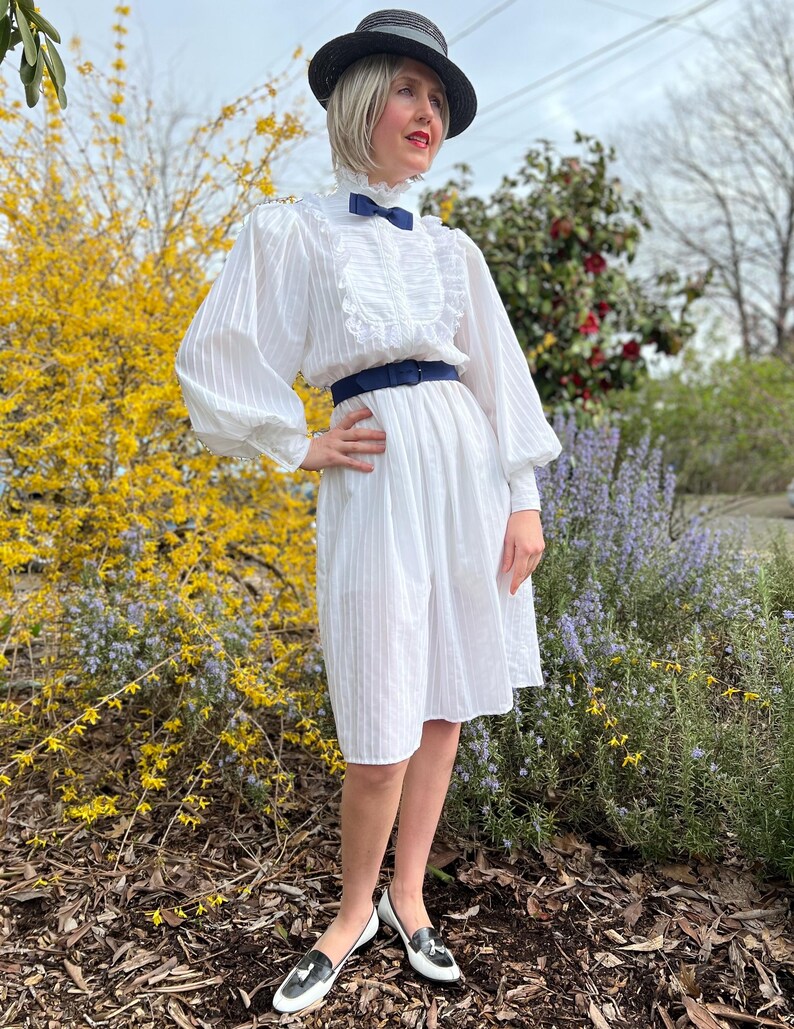 K&ouml;nnte beinhalten: Ein wei&szlig;es Kleid mit einem R&uuml;schenkragen und einer blauen Schleife. Das Kleid hat ein tailliertes Oberteil und einen vollen Rock. Das Model tr&auml;gt einen schwarzen Hut und schwarz-wei&szlig;e Schuhe.