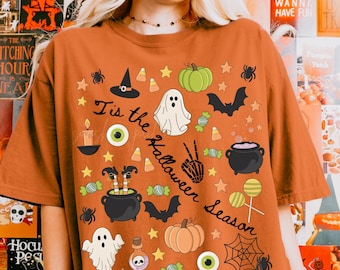 May include: Orange t-shirt with Halloween-themed graphics, including ghosts, pumpkins, bats, and the text "Tis the Halloween Season." The design features stars, candy corn, and a spiderweb.