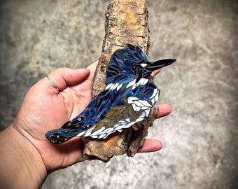 Könnte beinhalten: Ein Mosaik-Kunstwerk, das einen blau-weißen Eisvogel auf einem Ast zeigt. Der Vogel ist aus kleinen Glas- und Fliesenstückchen gefertigt, was einen strukturierten und farbenfrohen Effekt erzeugt.