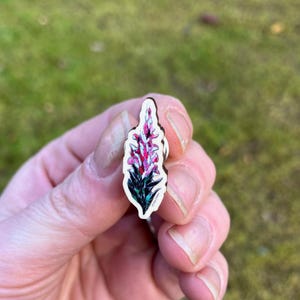 May include: A wooden pin featuring a colorful illustration of a foxglove flower. The flower has pink and purple blooms with green leaves. The pin is held in a hand against a blurred green background.
