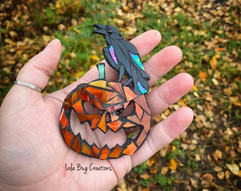 May include: A mosaic glass pumpkin with a black crow perched on top. The pumpkin is orange and has a scary face. The crow has iridescent blue and purple accents. The background is a blurry image of fall leaves.