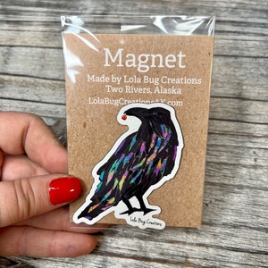 May include: A colorful raven magnet with iridescent feathers and a red berry in its beak. The magnet is packaged on a brown card with the text "Magnet" and "Made by Lola Bug Creations Two Rivers, Alaska LolaBugCreationsAK.com".