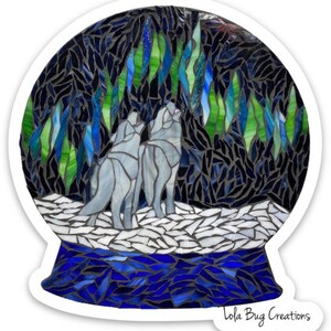 May include: A stained glass snow globe with two grey wolves howling at the aurora borealis, a green and blue light show in the night sky. The base of the snow globe is blue and white.
