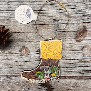 May include: A wooden ornament shaped like a boot, featuring a mushroom and foliage design. The top of the boot is yellow, with a brown lower section. A tag with the text "A.B. Creations" and a raven illustration is attached.