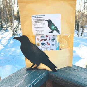 May include: A brown paper bag with a white label that says "Raven Forget-me-not DIY Mosaic Kit" and a picture of a raven with a forget-me-not flower. The bag is sitting on a wooden railing with a black metal raven silhouette in front of it.