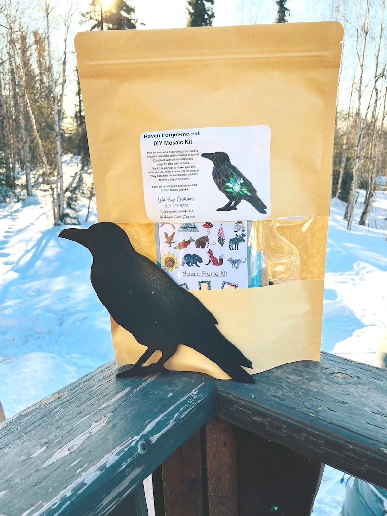 May include: A brown paper bag with a white label that says "Raven Forget-me-not DIY Mosaic Kit" and a picture of a raven with a forget-me-not flower. The bag is sitting on a wooden railing with a black metal raven silhouette in front of it.