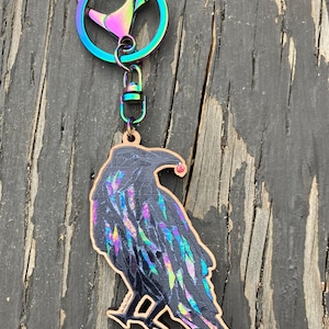 May include: A wooden raven keychain with a rainbow-colored metal clasp and ring. The raven is black with iridescent feather details and a small red berry. The keychain is on a weathered wooden surface.