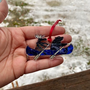 May include: A glass ornament depicting a moose and a bear in a blue canoe with white paddles. The ornament has a red ribbon attached for hanging.