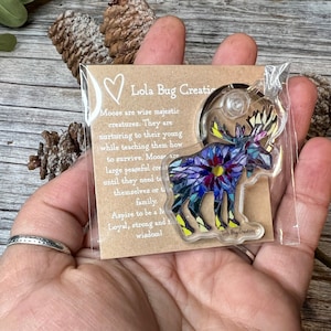 May include: A colorful mosaic moose keychain with a key ring. The keychain is packaged in a clear plastic bag with a brown card that reads "Lola Bug Creations" and includes a quote about moose.