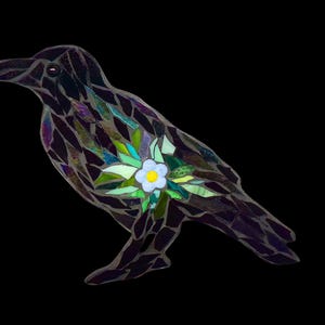 May include: A mosaic glass crow with iridescent purple and black feathers. A white flower with green leaves is embedded in the crow's chest.
