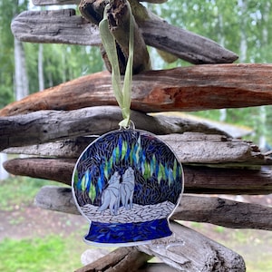 May include: A glass ornament shaped like a snow globe with a blue and green mosaic design. Two white wolves are howling at the moon inside the globe. The ornament is hanging from a green ribbon and has the text "Lola Bug Creations" at the bottom.