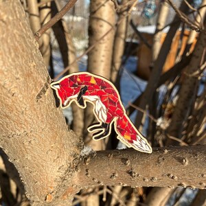 May include: A wooden fox-shaped ornament with a mosaic-like design in shades of red, orange, and white. The fox is perched on a tree branch, showcasing its detailed craftsmanship and artistic flair. The ornament has a black outline.