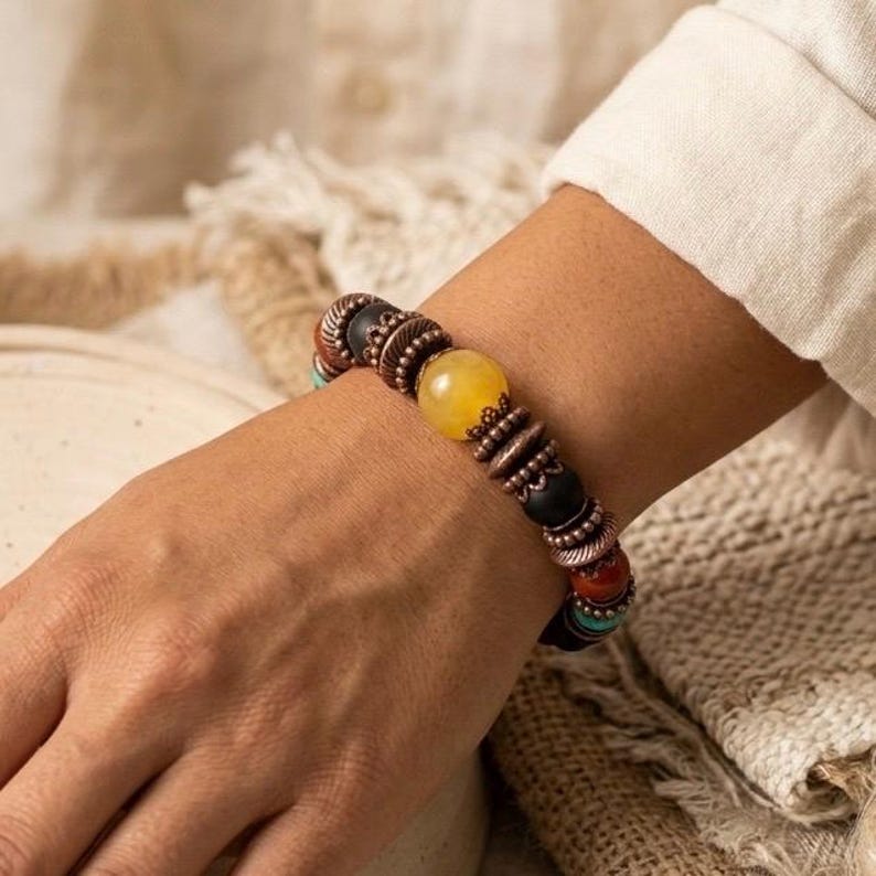 May include: A beaded bracelet featuring a large yellow bead, black beads, and smaller red, turquoise, and brown beads. The bracelet has copper-colored metal accents. The bracelet is on a wrist.