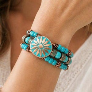 May include: A turquoise and copper-toned bracelet featuring three strands of turquoise beads. The center piece is a round copper medallion with a turquoise sunburst design. The bracelet is worn on a wrist.