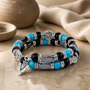 May include: A double-strand bracelet with black, grey, and turquoise beads, silver-toned metal spacers and decorative elements. A silver arrowhead charm hangs from the bracelet. The bracelet is displayed on a textured surface.