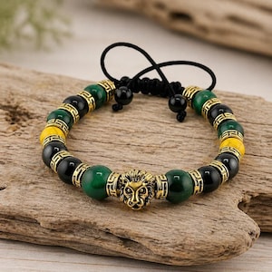 Op de afbeelding: Een kralenarmband met een gouden leeuwenkopbedel en groene, gele en zwarte kralen. De armband heeft een verstelbaar zwart koord. De kralen zijn geaccentueerd met goudkleurige metalen afstandhouders.