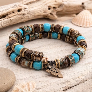 Könnte beinhalten: Ein Wickelarmband mit türkisfarbenen Perlen, dunkelbraunen Holzperlen und dekorativen Metallperlen. Ein bronzefarbener Pfeilanhänger verleiht diesem Accessoire im Bohème-Stil eine besondere Note.