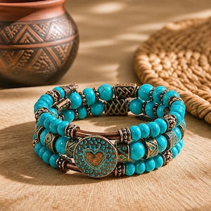May include: A three-strand bracelet with turquoise beads and antique bronze accents. The bracelet features a round, bronze charm with a heart design. The bracelet is displayed on a wooden surface.