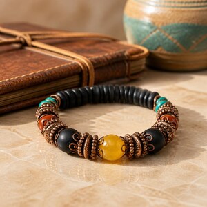 May include: A beaded bracelet featuring black, yellow, turquoise, and orange beads. The bracelet has copper-colored accents and a central yellow bead. The bracelet is on a light-colored surface.