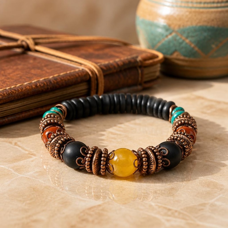 May include: A beaded bracelet featuring black, yellow, turquoise, and orange beads. The bracelet has copper-colored accents and a central yellow bead. The bracelet is on a light-colored surface.