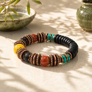May include: A beaded bracelet featuring a mix of colors and textures. The bracelet includes yellow, black, red, and turquoise beads, along with copper-colored spacers and a section of black, circular beads. The bracelet is displayed on a light-colored surface.