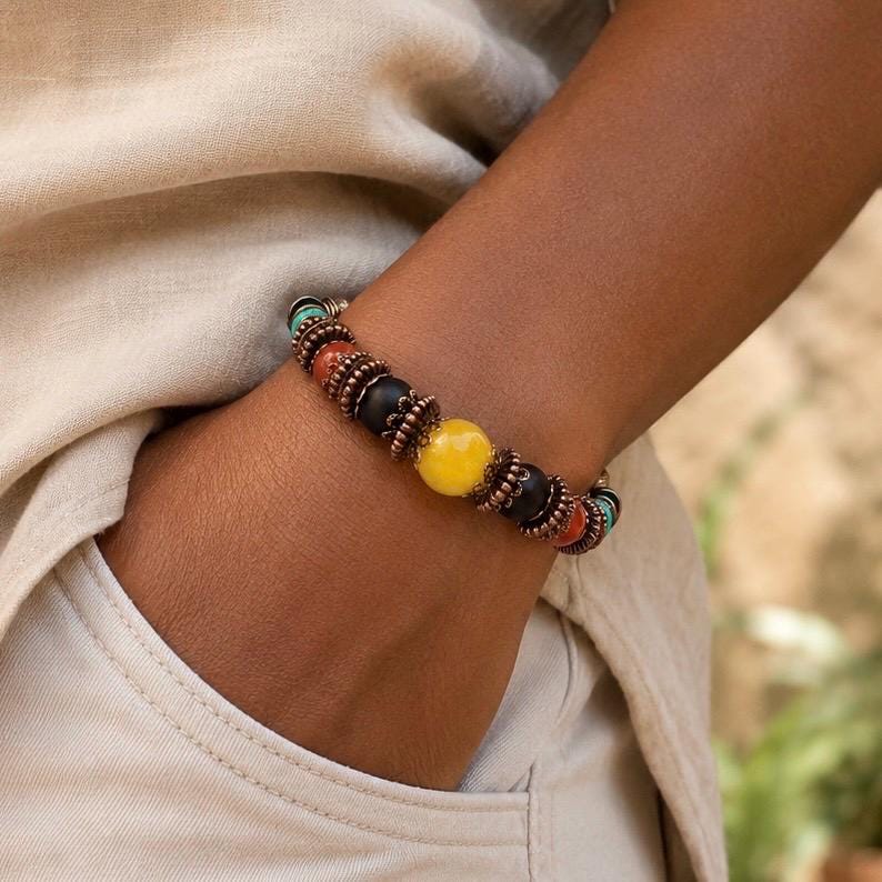 May include: A beaded bracelet featuring a mix of colorful beads, including yellow, black, red, and turquoise, strung together with copper-colored metal accents. The bracelet is worn on a wrist, showcasing its vibrant design.