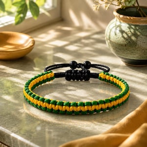 Può includere: Un bracciale in corda intrecciata con una fila centrale di nodi gialli affiancata da due file di nodi verdi. Il bracciale presenta una chiusura scorrevole nera regolabile con due piccole perline nere alle estremità dei cordini.
