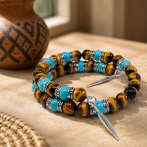 May include: Two wrap bracelets featuring tiger eye beads, turquoise beads, and silver-coloured accents. Each bracelet has a silver feather charm. The bracelets are displayed on a light brown surface.