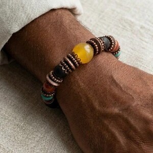 May include: A beaded bracelet featuring a large yellow bead, black, orange, and turquoise beads, and copper-colored metal accents. The bracelet is worn on a wrist, showcasing the colorful design and textures.