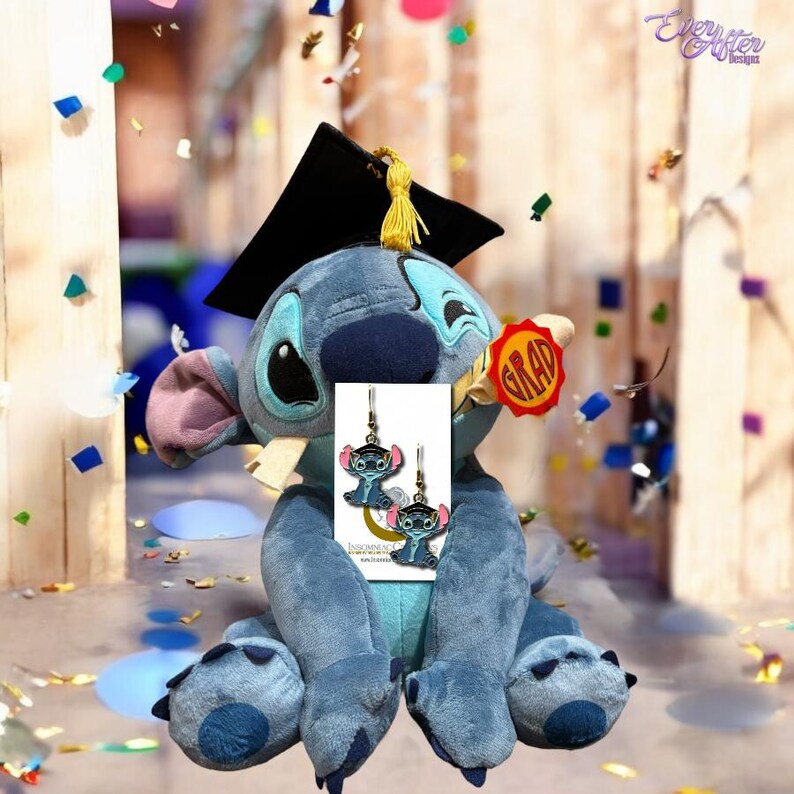 May include: Blue Stitch plush toy wearing a graduation cap and holding a diploma with the word 'GRAD' on it. The Stitch plush toy is holding a card with two pairs of blue Stitch earrings on it.