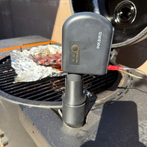 Puede incluir: Un controlador de termómetro digital Inkbird negro montado en una ventilación de parrilla. El dispositivo cuenta con una luz de estado circular, una pequeña luz indicadora etiquetada WiFi STATUS y un cable de sonda rojo en el lateral.