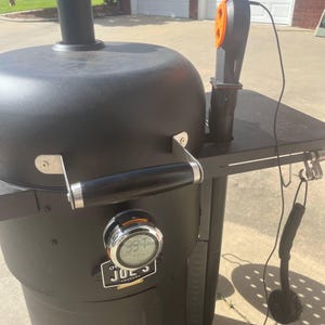 May include: Black smoker grill with a digital thermometer displaying 397 degrees. The grill has a side shelf, a handle, and wheels for easy movement. An orange and black temperature probe is attached.