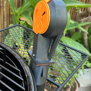 May include: A black ChefsTemp grill thermometer with an orange dial and accents. The thermometer is attached to a black metal grill with a wire rack. The background features green plants and a bamboo fence.