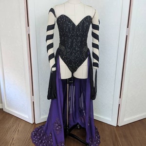 May include: A black corset with a sweetheart neckline and a purple skirt with a sheer overlay. The corset is embellished with black embroidery and rhinestones. The skirt has a beaded trim.