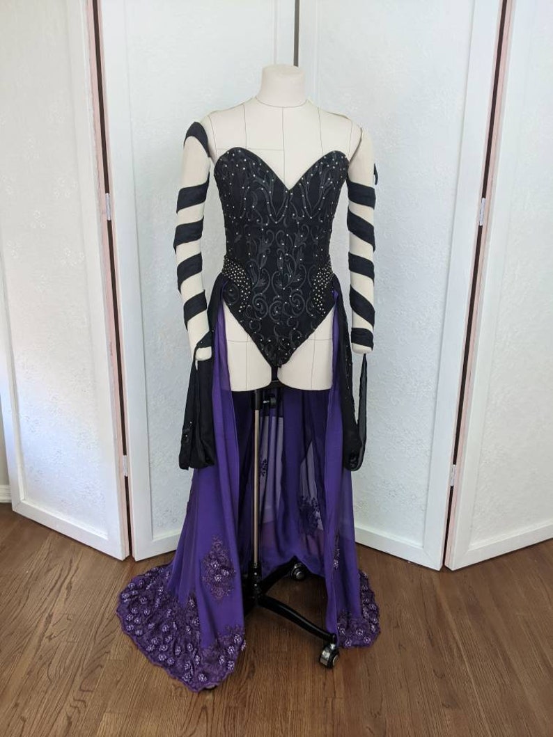 May include: A black corset with a sweetheart neckline and a purple skirt with a sheer overlay. The corset is embellished with black embroidery and rhinestones. The skirt has a beaded trim.