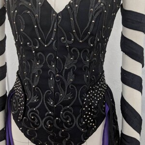 May include: A black corset with a sweetheart neckline and a floral design. The corset is embellished with rhinestones and has a purple ribbon attached to the back.