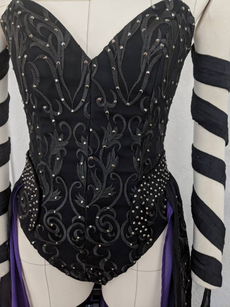 May include: A black corset with a sweetheart neckline and a floral design. The corset is embellished with rhinestones and has a purple ribbon attached to the back.