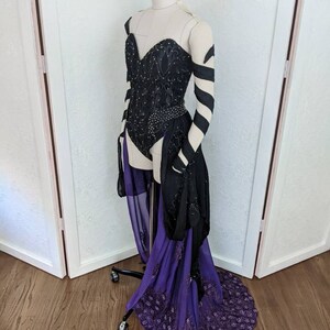 May include: A black and purple costume with a sweetheart neckline, long sleeves, and a flowing skirt. The bodice is embellished with sequins and the skirt has a lace trim.