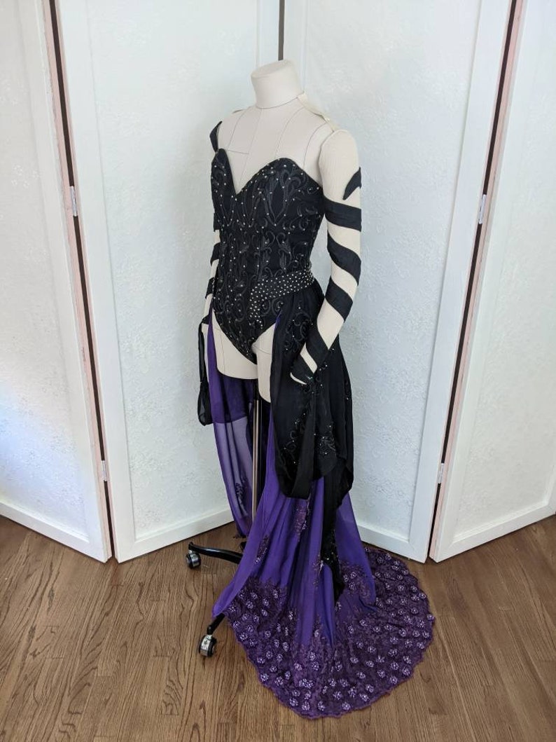 May include: A black and purple costume with a sweetheart neckline, long sleeves, and a flowing skirt. The bodice is embellished with sequins and the skirt has a lace trim.