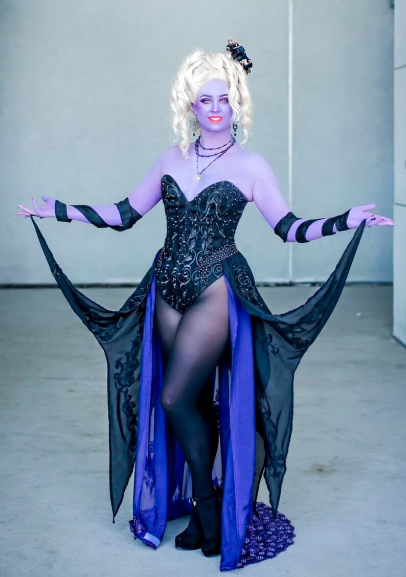 May include: A woman in a purple and black costume with a long flowing skirt and a black corset. She is wearing a black wig and has a crown on her head.