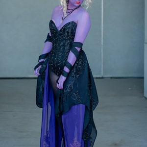 May include: A woman in a black and purple dress with a black corset and a purple sheer overlay. She has purple makeup and a black headband with a small crown.