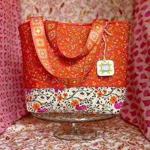 May include: A quilted tote bag with a vibrant floral pattern in shades of orange and pink. The bag features a contrasting floral border and patterned straps. A tag with the text "Lavender Haze Press" is attached.
