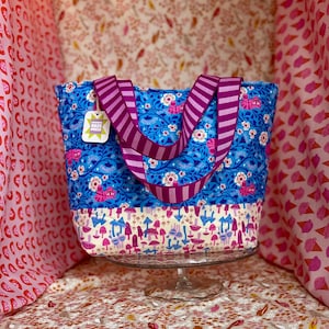 Op de afbeelding: Een blauwe draagtas met een bloemenpatroon en gestreepte paarse handvatten. De tas heeft een crèmekleurig onderste paneel met een paddenstoelontwerp. Een label met de tekst "RAZE PRESS" hangt aan het handvat.