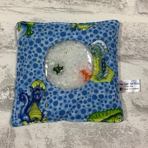 May include: Blue sensory bag with a clear window filled with white beads. The bag has a green and blue dragon and turtle design. The bag has a tag that says "Sew Cool for Kids I-Spy Bag Find us on Facebook".