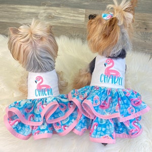 May include: Two small dogs wearing matching blue and pink dresses with a flamingo print. The dresses have a white bodice with a pink flamingo and the name "Chichi" and "Charli" embroidered on them.