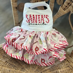 May include: A white bib with the text "SANTA'S favorite" in red embroidery. The bib is attached to a pink and white Christmas-themed dress with red striped trim and a tiered skirt. The dress features Santa Claus and candy cane patterns.