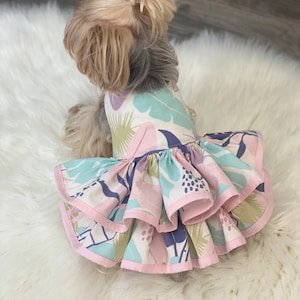 May include: A small dog wearing a floral print dress. The dress features a white background with pastel-colored leaves and a ruffled skirt with pink trim. The dog is sitting on a white, fluffy surface.